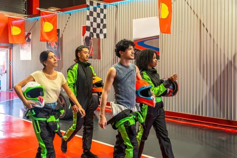 Group of diverse gen z friends holding racing helmets and walking in an indoor karting facility,