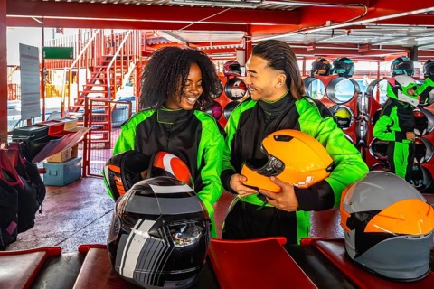 Happy young multiracial couple standing in an indoor karting track area, holding helmets, and
