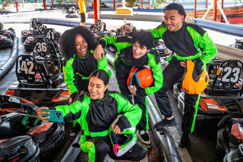 Diverse group of friends in racing suits and helmets smiling for a selfie at a karting track,