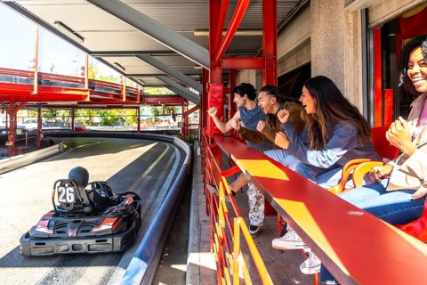Group of excited diverse friends cheering for a go kart speeding on an indoor multi level track,
