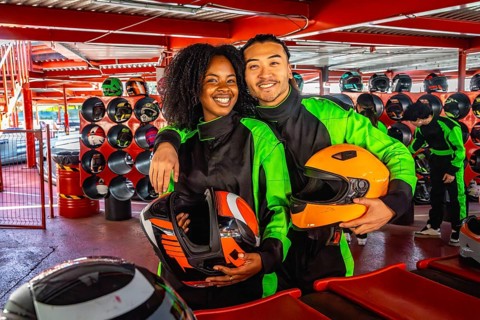 Young couple in racing suits with helmets, smiling at the camera on an indoor go kart track, ready