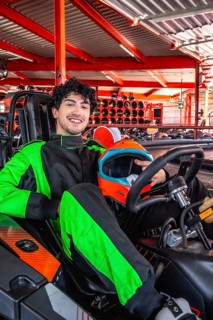 Young man in green and black racing suit smiles holding orange and blue helmet while seated in a go