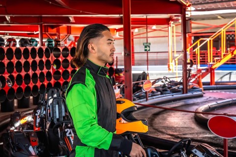 Young man wearing a racing suit standing with an orange helmet by a go kart, preparing to drive on