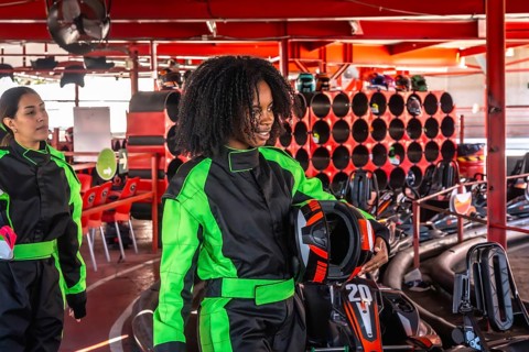 Two cheerful young women in racing suits and holding helmets preparing to race go karts at an