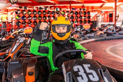 Asian man in racing suit and yellow helmet sits in a go kart on track, smiling and pumping fist in