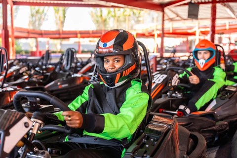 Young woman smiling in a black and neon green racing suit and orange helmet, sitting in a go kart