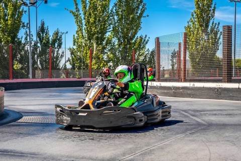 Drivers are speeding go karts around a curve on an outdoor asphalt track, wearing protective