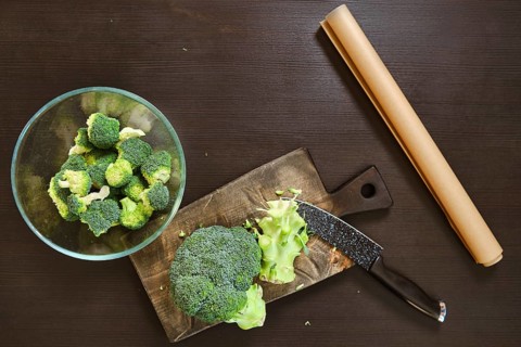 Broccoli is being cut on a wooden board. A bowl of chopped broccoli sits nearby. The kitchen is