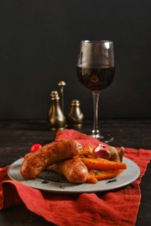 Sausages made with natural casings are placed on a plate with colorful vegetables. A glass of red