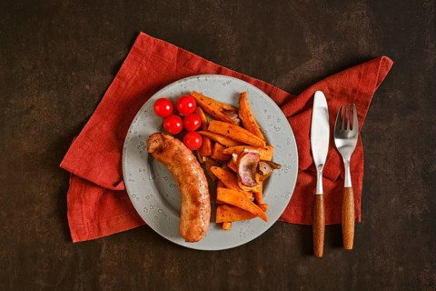 A plate holds natural casing sausages alongside roasted sweet potatoes and cherry tomatoes. The
