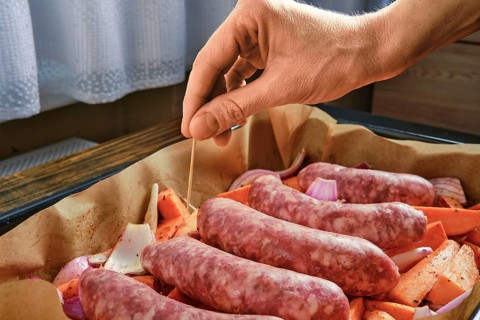 A hand is pierces a toothpick into natural casing sausages arranged on a tray with chopped