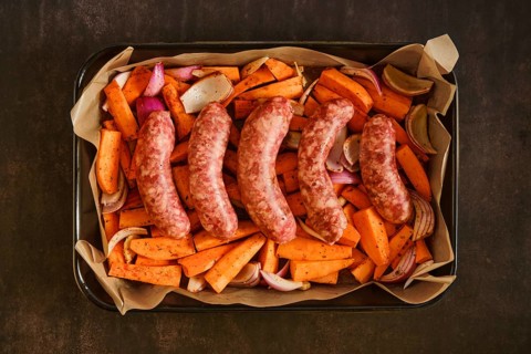 Raw sausages are placed on a baking tray with sliced sweet potatoes, onions, and garlic. The scene