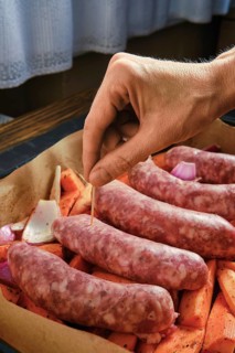 A hand is placing a toothpick into natural casing sausages that rest on a bed of sweet potatoes and