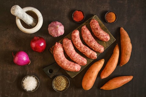 Natural casing sausages on a wooden board alongside sweet potatoes, garlic, onions, and various