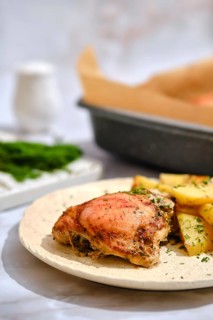 A plate holds a crispy chicken thigh next to golden roasted potatoes. Fresh green herbs are