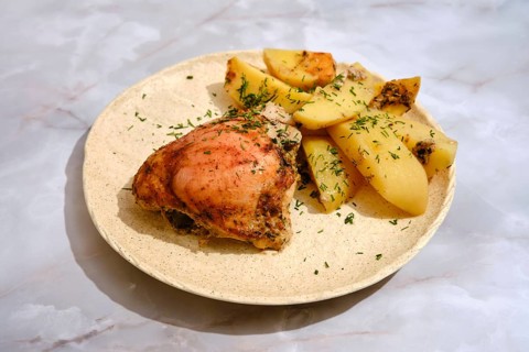 A portion of baked chicken is placed on a plate alongside cut potatoes. The chicken has a bit of