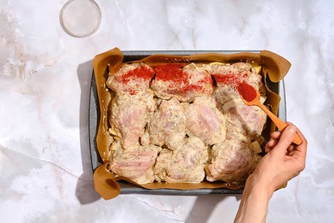 A person spreads smoked paprika over chicken thighs placed in a baking tray lined with parchment