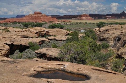 Wide rocky landscape with natural water basins and large red rocks in the background under a cloud