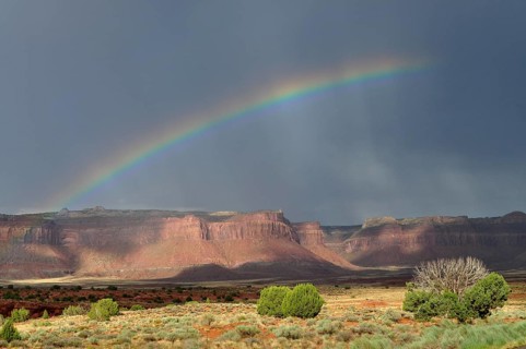Bright rainbow stretches across a dramatic desert landscape with rocks and threatening sky,