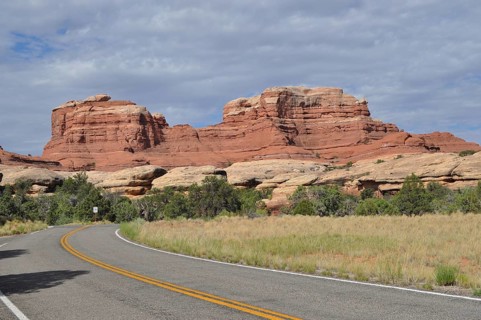 A winding country road runs in front of an impressive red rock formation under cloudy skies,