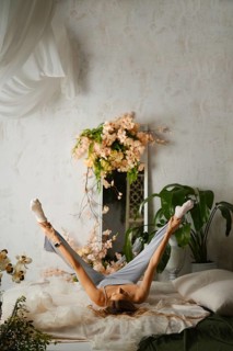 A woman is lying on a bed and stretching her legs in the air. The room has flowers and green plants