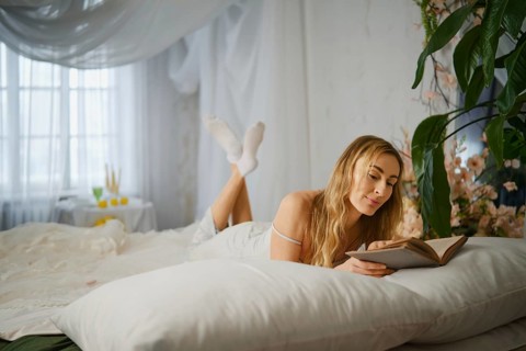 A woman is lying on a bed, reading a book with a relaxed pose. The room is bright and has plants