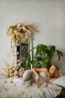 A woman relaxes on a bed while reading a book in a sunny room. She is surrounded by plants and