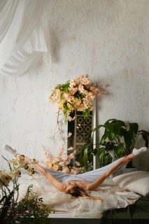 Woman in pajamas stretches on a bed in a bright room filled with plants and flowers. The stretching