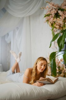 Reading a book while lying on a bed in a room with flowers and a plant during afternoon light