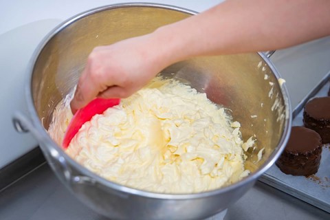 A hand stirs buttercream in a large metal bowl with dough spatula, vegan cake production,