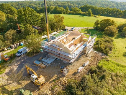 Construction site with timber structure and excavator, surrounded by green landscape and a crane,