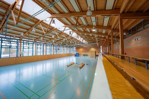Spacious, bright sports hall with visible wooden roof and large window fronts along the walls,