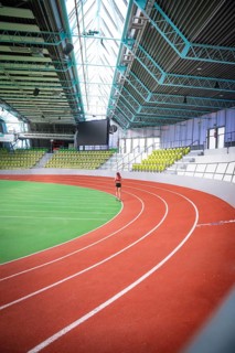 A person runs on an oval, red-green running track of a spacious indoor sports hall, middle distance