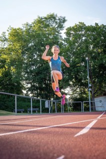 A person wearing blue sportswear jumps with raised arm on the running track in sunshine, middle
