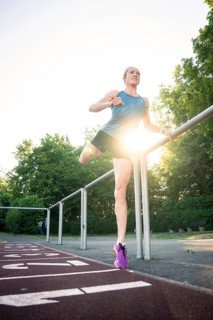 Woman stretching on a track in sunlight, holding onto the railing, surrounded by trees, middle