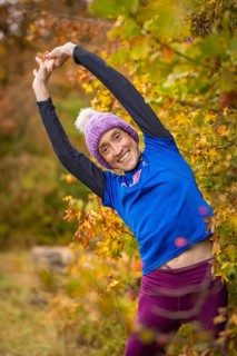 Woman doing stretching exercises in autumn forest, smiling happily surrounded by colorful leaves,