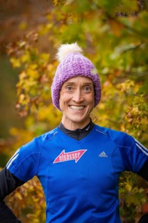 Person wearing blue clothes and purple cap smiling in autumn surroundings, middle distance runner