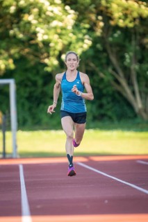 Person running on a red track surrounded by green trees and bright sunlight, middle distance runner