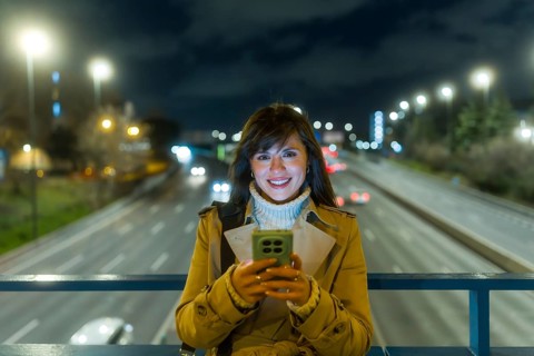 Young woman standing on an urban bridge at night, smiling while reading messages on her mobile