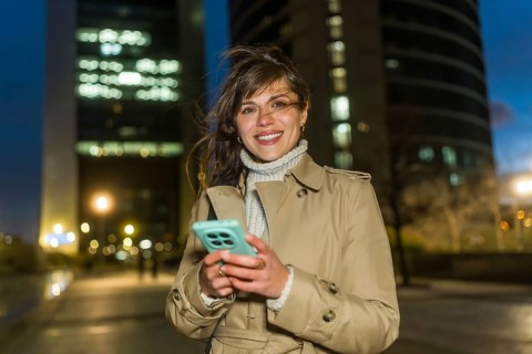 Young woman in a trench coat stands on a lit city street at dusk, smiling at the camera while