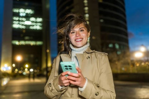 Young woman smiling, holding her smartphone, and looking at the camera outdoors in a modern city