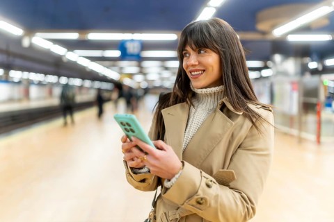 Woman standing on a train station platform, smiling and looking away while sending messages or