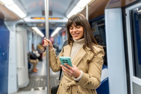 Young woman standing in a subway car, smiling at her smartphone screen during the commute, browsing