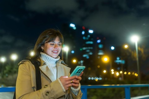 Young woman smiling as she types on her smartphone on a city street at night, illuminated by bokeh