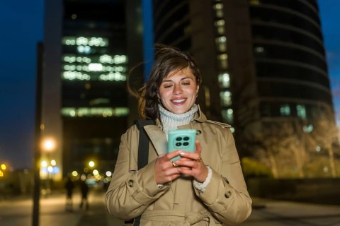 Young woman smiling while walking through the city at night, engaging with her mobile phone and
