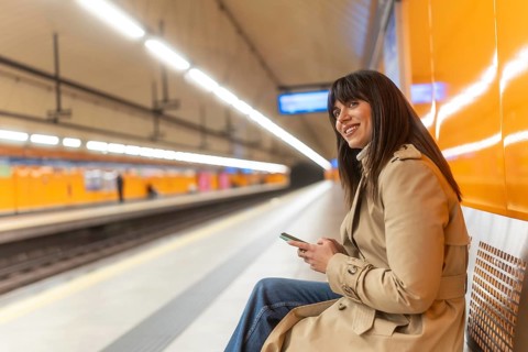 Young woman commuting, checking her mobile phone and smiling while sitting on a bench at an empty