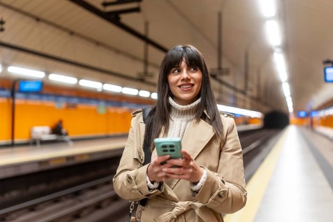 Young woman in a trench coat and sweater smiling while using her smartphone and waiting for a train