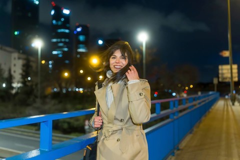 Young woman in a trench coat smiling, touching her hair, and looking at the camera while standing