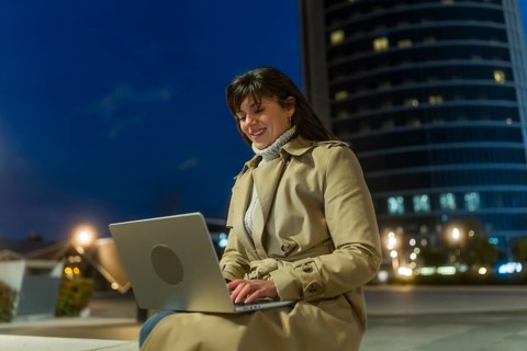 Woman in a trench coat smiling and typing on a laptop, connecting to the internet and managing her