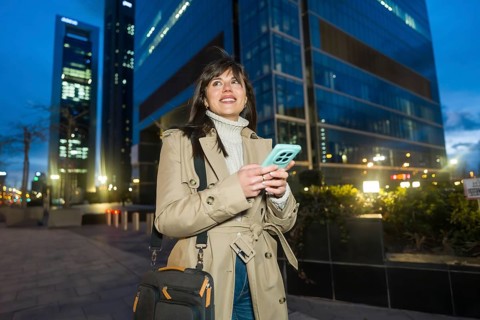 Young professional woman walking along a city sidewalk at dusk, holding a smartphone and carrying a